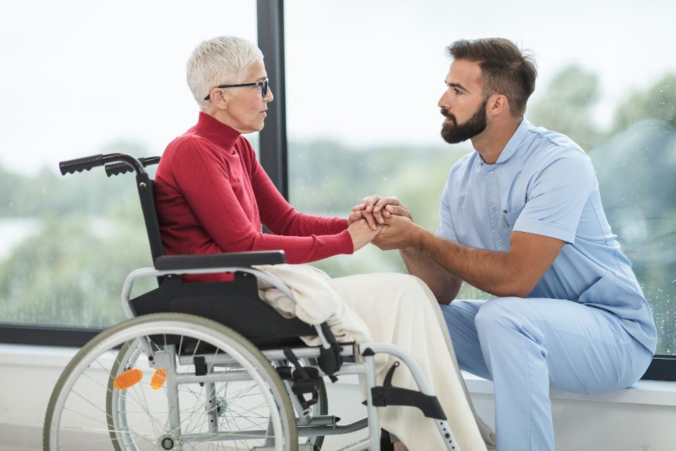 A caregiver kneels beside a smiling senior woman in a wheelchair and holds her hands